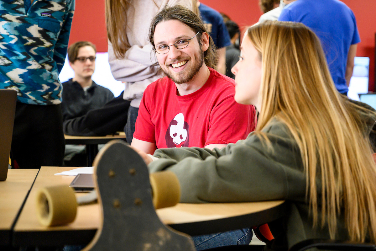 Two students sit at a table. One is smiling at the other. 